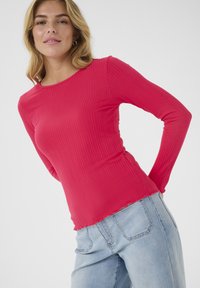 Ribbed long-sleeve top in bright pink, featuring a rounded neckline and uneven hem, paired with light blue denim jeans.