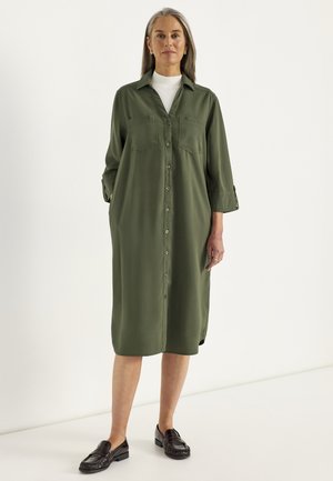 Kvinde med gråt hår iført en olivengrøn midi-kjole med knapper over en hvid top og sorte loafers, stående mod en ensfarvet baggrund.