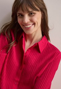Camicia aderente di un vivace colore rosa, in tessuto texturizzato con strisce verticali, con chiusura a bottoni e colletto, presenta una tasca sul petto e maniche lunghe.