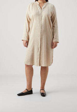 Kvinde iført en beige knap-ned-langærmet knælængde kjole med sorte flade Mary Jane-sko, stående mod en ensfarvet baggrund.