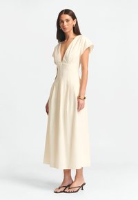 Ivory mekko, jossa on syvä V-pääntie, hihattomat hihat ja istuva vyötärö, jotka laskeutuvat säären puoliväliin. Sujuva kangas, jossa on hienovaraisia laskostusdetaljeja.