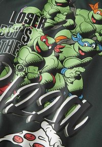 Melnā T-krekla ar grafiku, kurā redzamas četras zaļas bruņurupucis dažādās pozās, ko papildina krāsaini akcenti un teksts, kas vēsta: "LOSER BUY $ THE PIZZA."
