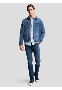 Mann trägt blaue Jeansjacke, hellblaues Hemd mit Knöpfen, blaue Jeans und marineblaue Turnschuhe, geht mit einer Hand in der Tasche.