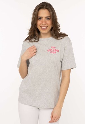 Lächelnde Frau mit lockerem braunem Haar, die ein hellgraues "Feel Good Club"-T-Shirt und weiße Hose trägt und den Kragen des T-Shirts hält.