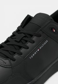 Black sneaker with smooth synthetic upper, round toe, flat laces, and logo detailing. Features a textured panel and contrasting accents.