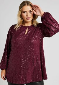 Burgundy paljettopp med långa, puffiga ärmar och detalj med nyckelhål i halsringningen. Strukturerad, glänsande yta med en avslappnad passform vid midjan.