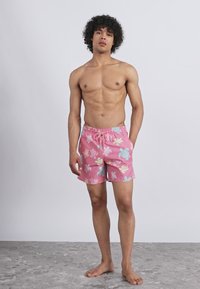 Tânăr stând desculț, purtând șorturi de baie roz cu modele pastel de broaște țestoase, pe un fundal simplu deschis.