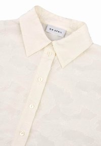 Camicia color crema con colletto realizzata in tessuto testurizzato, caratterizzata da un delicato motivo floreale e sei bottoni lungo la parte frontale.