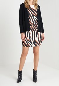 Un cardigan noir sur une robe blanche avec un motif abstrait orange et noir. Mannequin portant des bottines noires. Design à longueur de sol.