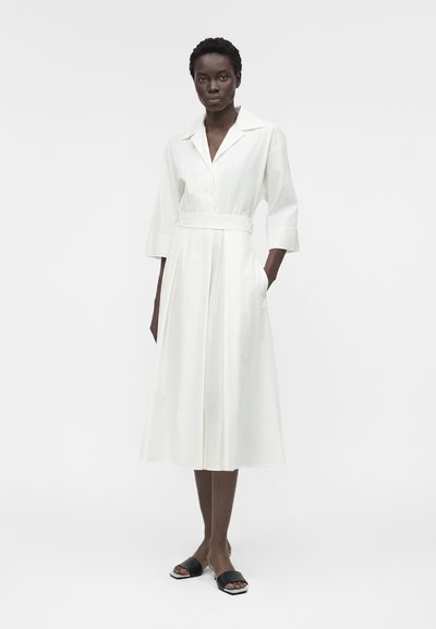 Γυναίκα με κοντά μαλλιά που φοράει λευκό μεσάτο φόρεμα με ζώνη και μανίκια τρία τέταρτα, καθώς και μαύρα πέδιλα με ανοιχτά δάχτυλα, στέκεται μπροστά σε απλό φόντο.