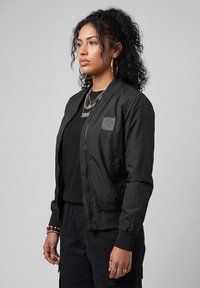 Woman with curly hair wearing a black bomber jacket, black shirt, layered necklaces, hoop earrings, and multiple rings and bracelets stands sideways.