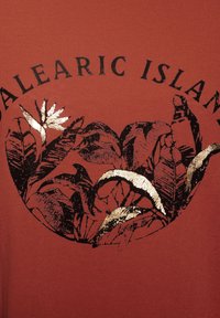 Rustfarbiges T-Shirt mit einem grafischen Design von schwarzen Blättern und einer Blume mit goldenen Akzenten, der Text "BALEARISCHE INSELN" steht über dem Bild.