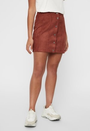Femme portant une mini-jupe boutonnée couleur rouille, un haut blanc et des baskets blanches épaisses, debout devant un fond uni.