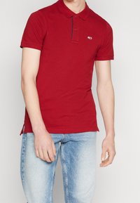 Červené bavlněné polo tričko s límečkem a dvoubarevným knoflíkovým zapínáním. Obsahuje modré akcenty, krátké rukávy a postranní rozparky na lemu.