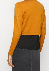 Suéter corto de tejido de punto en color dorado-naranja con mangas largas, combinado con una blusa negra de capas y una falda de denim gris oscuro.
