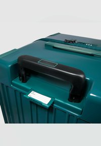 Turkooizen harde koffer met een zwart handvat, een houder voor een adreslabel en een combinatieslot. Ribbelstructuur met een gladde afwerking.