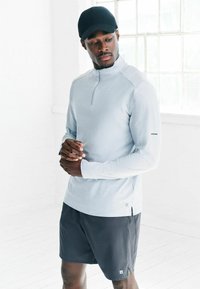 Vīrietis, kurš valkā melnu cepuri, gaiši pelēku garām piedurknēm ar rāvējslēdzēju, un tumši pelēkas šortus, stāv iekštelpās blakus lieliem logiem uz baltiem sienām.