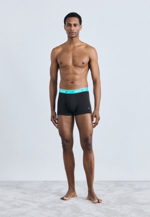 Schwarze Boxershorts mit einem hellblauen Nike-Bund, die eine schmale Passform und Baumwollmaterial aufweisen. Das Modell steht barfuß vor einem neutralen Hintergrund.