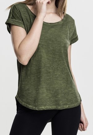 Frau trägt ein kurzärmeliges olivgrünes T-Shirt und schwarze Hose, eine Hand nahe am Gesicht, die andere auf der Hüfte.
