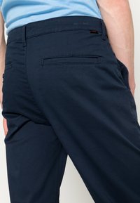 Dunkelblaue Chinos mit glatter Textur, ausgestattet mit zwei Gesäßtaschen und einem geraden Bein Design. Sichtbare Nähte und Etikettendetail.