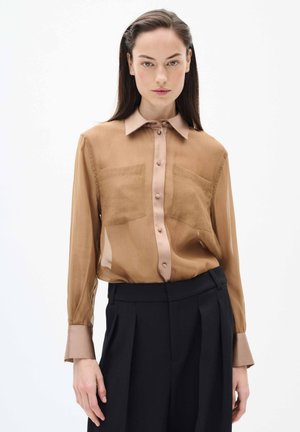 Lichtbruine, transparante blouse met een lichte kraag en zes knopen, voorzien van twee voortassen en getailleerde mouwen, gecombineerd met donkere broeken.