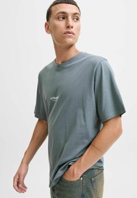 Grau meliertes Baumwoll-T-Shirt mit kurzen Ärmeln und Rundhalsausschnitt, mit einem kleinen weißen Logo auf der Brust, getragen mit blauen Jeans.