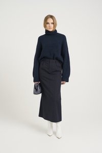 Granatowy sweter z golfem i szerokimi rękawami, zestawiony ze spódnicą ołówkową w paski. Modelka trzyma małą granatową torbę i nosi białe kozaki na obcasie.