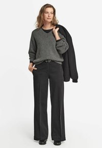 Szary sweter z dzianiny z dekoltem w serek, w połączeniu z czarnymi spodniami o szerokich nogawkach i czarną marynarką zarzuconą na jedno ramię. Gładka faktura, luźny krój.