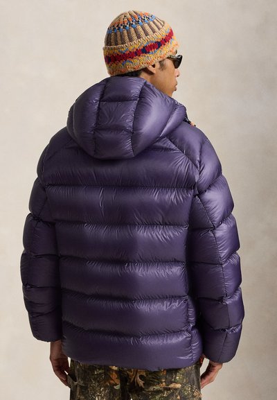 Man wearing a purple puffer jacket with a hood, a multicolored knit beanie, sunglasses, and camouflage pants facing away.