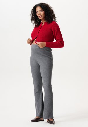 Femme aux cheveux bouclés portant un pull rouge boutonné et un pantalon gris côtelé taille haute, debout devant un fond uni.