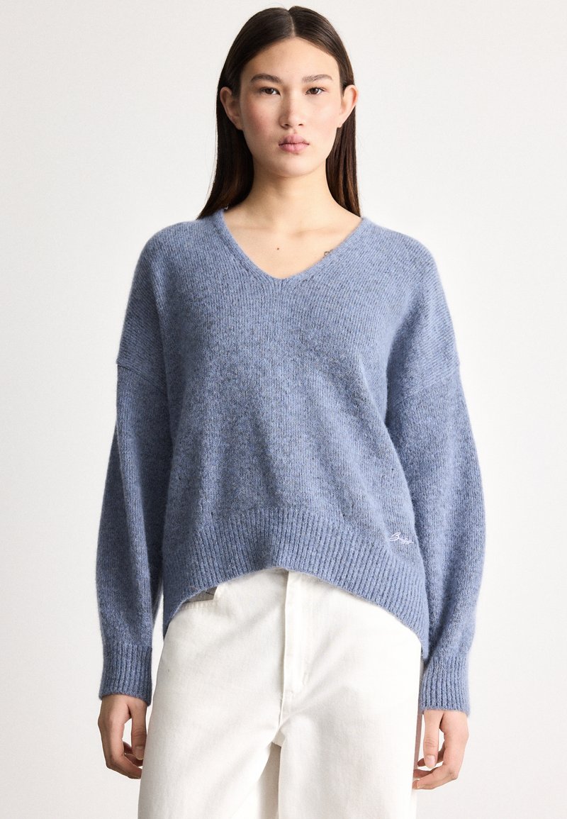 Μπλε, άνετο πλεκτό πουλόβερ με V-λαιμόκοψη, ριμπ τελείωμα και φαρδιά μανίκια. Διαθέτει ελαφρώς ανοιχτό μήκος εμπρός-πίσω και κεντητό διακοσμητικό στην κάτω πλευρά.