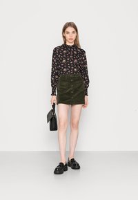 Blusa preta de manga longa com padrão floral e punhos franzidos, combinada com uma saia mini em corduroy verde escuro, com botões frontais e dois bolsos. Bolsa e sapatos pretos.