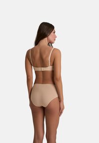 Beige draadloze beha met verstelbare banden en haaksluiting, gedragen met bijpassende hooggetailleerde onderbroek, die een gladde, naadloze textuur laat zien.