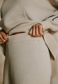 Beež värvusega kudumist kampsun esitasku ja soonikkoes varrukaäärtega, koos sobivate soonikkoes vöökohaga pükstega. Pehme tekstuur ja õmblusteta disain.