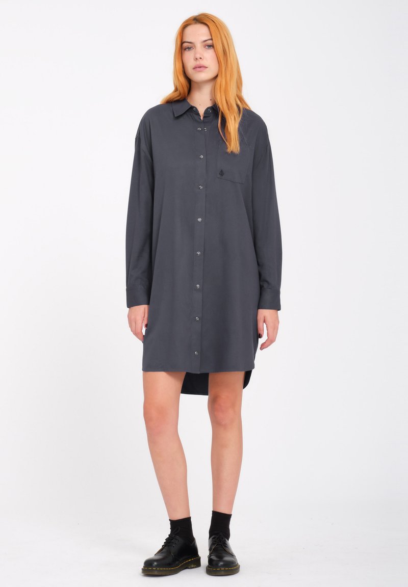 Donkergrijze langekjesshirt met knoopsluiting, enkele borstzak, gebogen zoom en lichtgewicht stof. Gemaakt met zwarte enkellaarzen.