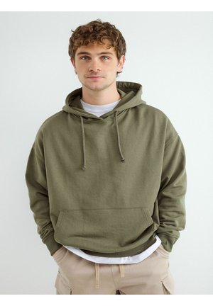 Młody mężczyzna o kręconych włosach ubrany w oliwkowo-zieloną bluzę z kapturem na białej koszuli i beżowych spodniach, stojący na jednolitym białym tle.