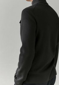 Dark sweater with a high collar, smooth texture, and fitted design. Features a subtle sheen and a pocket detail on the left side.