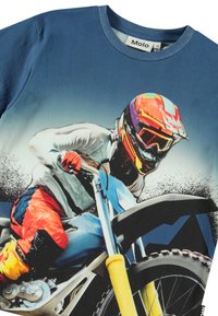 Modrá trička s motocrossovým motivem, na kterém je jezdec v barevném oblečení. Měkká tkanina, kulatý výstřih, krátké rukávy a živý design.