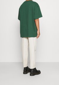 T-shirt oversize vert foncé avec un col rond, assorti à un pantalon beige clair et des bottines noires. Coutures visibles et coupe décontractée.