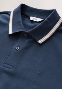 Tumesinine polo-särk beežide triibulise kontrastkraega. Omadused: kahe nupuga plastron ja väike tikitud krooni logo.