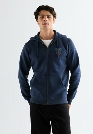 Młody mężczyzna w granatowej bluzie z kapturem na zamek, z przednimi kieszeniami i logo na piersi, w czarnych spodniach, stojący na jednolitym tle.