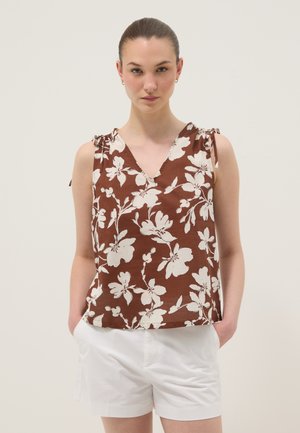 Vrouw draagt een bruine mouwloze bloemen top met schouderstrikjes en witte korte broek, staat met handen in zakken tegen een effen achtergrond.