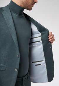 Ciemnozielony garnitur z teksturowanej tkaniny, z jasnoniebieską podszewką i wieloma kieszeniami wewnętrznymi. Smukła sylwetka z subtelnymi akcentami.