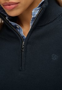 Marineblauwe gebreide pullover met een kraag met halve rits en ribbeltextuur. Heeft een lichtblauwe geruite overhemdkraag eronder en een geborduurd logo.