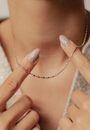 Goldene Perlenketten-Halskette, gehalten von zwei Fingern mit hellgrau polierten Nägeln vor nacktem Hals und beiger Kleidung.