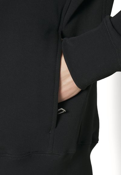 Svart hettegenser med teksturert stoff, med sidelomme og synlig varemerke. Ermmene er litt tettsittende for en komfortabel avslutning.