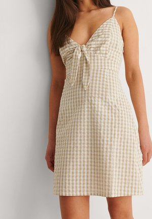 Kobieta ubrana w beżowo-białą sukienkę w kratę bez rękawów z wiązaniem z przodu, stojąca na tle jednolitego jasnego tła.