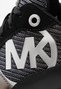 Baskets noires avec une tige texturée, des accents argentés, un logo "MK" bien visible et un détail circulaire en métal argenté.