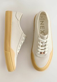 Paire de baskets beiges en toile avec semelles en gomme et lacets blancs, une chaussure vue de côté, l’autre vue de dessus montrant la taille et la marque.
