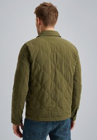Olivgrüne, gesteppte Jacke mit einem strukturierten Wellenmuster, Druckknöpfen und Stehkragen. Kombiniert mit blauen Jeans.